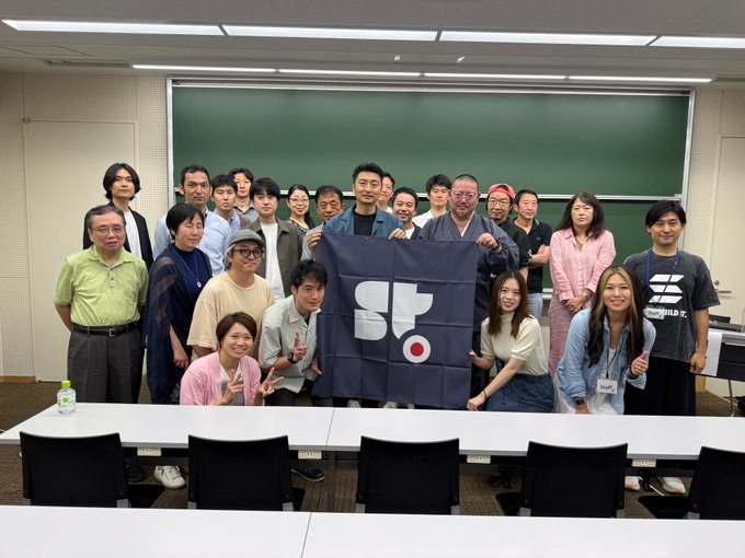 A group of approximately 20 people poses in a University of Tokyo classroom, with two individuals holding up a dark blue banner featuring a stylized "SF" logo and a red circle, likely from Stable Corp/Studio. The gathering relates to the 2026 Blockchain Application Practice Program outcomes presentation, where attendees reviewed projects on topics including on-chainization of traditional crafts and cross-border remittances to Southeast Asia.