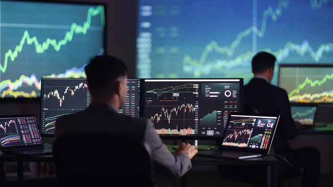 Two men in suits sit at a desk analyzing multiple monitors displaying green candlestick stock charts in a dark trading environment.