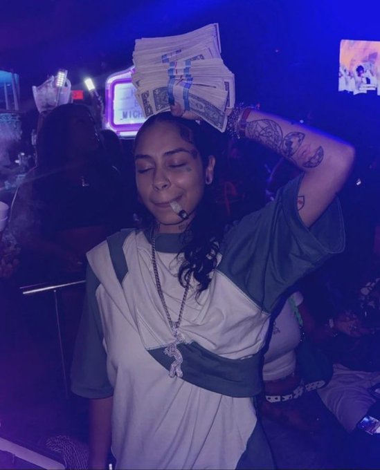 A woman smiling in a dimly lit club setting, holding a stack of US dollar bills above her head, wearing a white top and multiple necklaces.