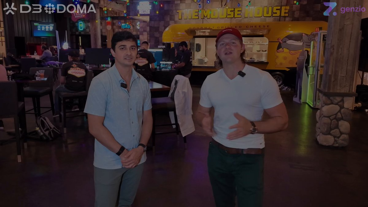 The image shows two men standing next to each other in a room, with one man wearing a white shirt and the other wearing a red cap. They are both smiling and appear to be engaged in conversation or discussing something related to their surroundings. The background of the image features a yellow truck parked behind them, suggesting that they might be at an event or gathering. There is also a TV visible in the scene, which could be used for presentations or announcements during the event.