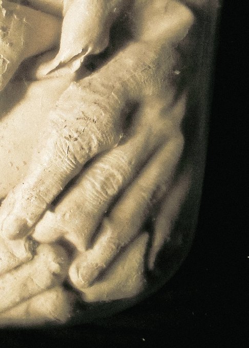 A close-up black-and-white photograph showing two elderly, wrinkled hands clasped together, with one hand resting gently atop the other in a tender gesture.