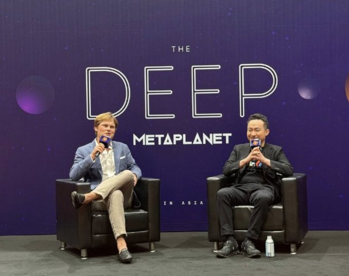 Two men sit in black armchairs on a stage holding microphones at 'THE DEEP METAPLANET IN ASIA' conference event, with one wearing a light blue blazer and the other in a black suit.