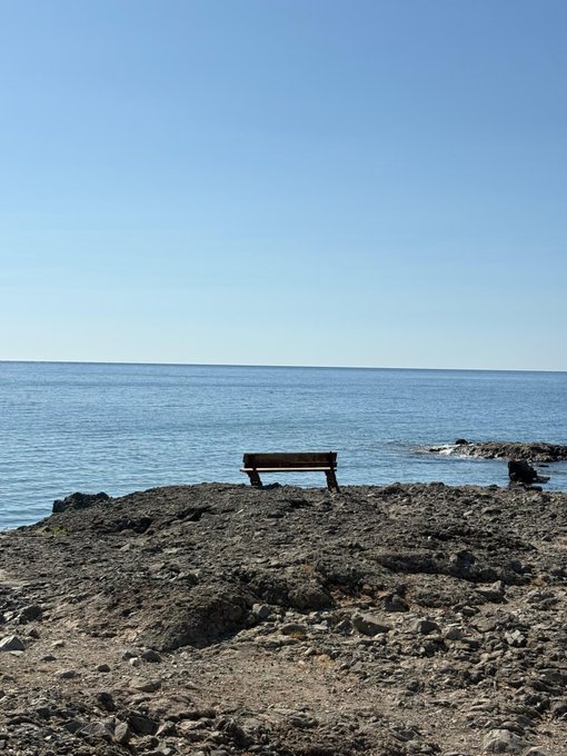 A lone wooden bench sits on a rocky shore overlooking the calm sea under a clear blue sky, symbolizing a moment of quiet reflection or strategic observation—fitting for a post about patiently tracking decentralized (DC) project roles over the next 5-6 months, including @base, @RialoHQ, @ritualnet, and @axisrobotics.