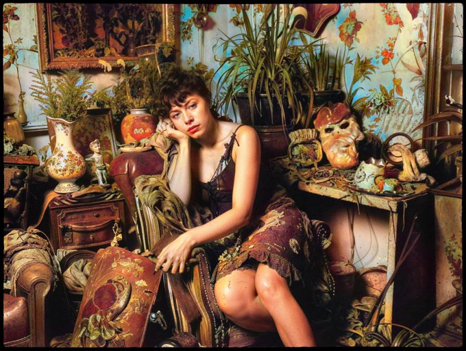 A photograph of a woman with short dark hair sitting in a richly decorated, cluttered room filled with antique vases, masks, and floral wallpaper.