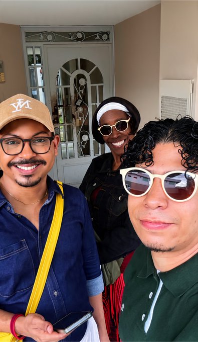 Selfie of three smiling friends wearing glasses and caps, standing in front of a doorway.