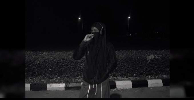 A black-and-white image of a person wearing dark clothing and a face covering, standing outdoors at night with streetlights in the background.