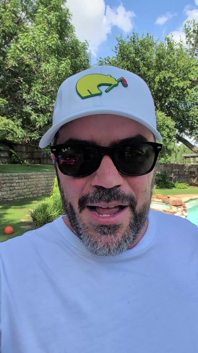 The image features a man wearing sunglasses, a white baseball cap with a green logo, and a t-shirt. The man has a beard and appears to be smiling for the camera. In the background, there are two golf balls visible on the grassy area near trees. The caption of the image reads "Video Actualización de MSTR y Bitcoin. Además, mi último análisis de la situación de AI y lo que pasó hoy con Apple and su nueva inteligencia artificial". This suggests that there might be a connection between the man's attire and the recent release of a new video game or a post about Bitcoin.