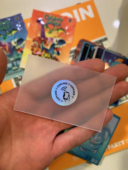 A hand holds a transparent card featuring a "CUPCAKE ©CUPCAKE" sticker, with colorful Claynosaurz packaging and merchandise visible in the background, highlighting the brand's journey from small-scale collectibles to an Amazon Prime launch.