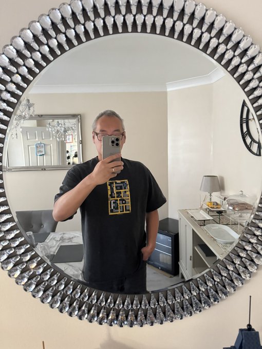 A man with glasses takes a mirror photo of his reflection in an ornately bordered round mirror while wearing a black t-shirt with a graphic design.