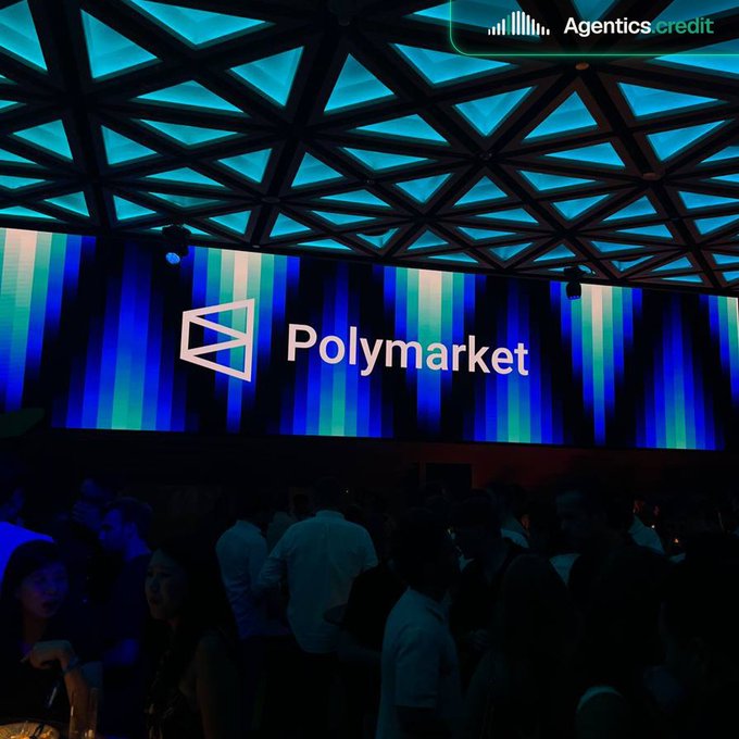 A large illuminated 'Polymarket' logo displayed on a screen at an event with a geometric ceiling, with a crowd gathered below and an 'Agentics.credit' watermark.