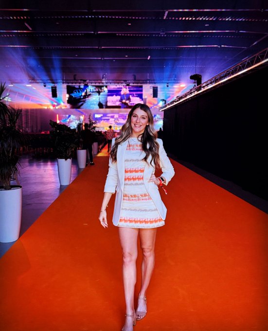 The image features a woman wearing an orange dress, standing on a red carpet with her arms outstretched as if she's posing for a photo. The background includes a stage or event setup with lights and potted plants, suggesting that this could be a formal gathering or performance. The caption "Great first day at @BTCPrague" implies that the woman is attending an important event in the city of Berlin, which has been described as a great first day for her.