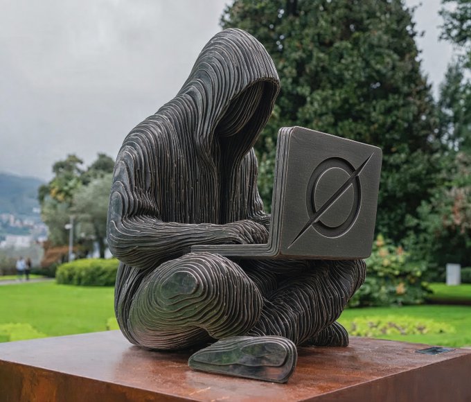 A textured metal sculpture of a hooded figure working on a laptop sits outdoors on a rust-colored pedestal, with a logo embossed on the laptop's lid, set against a green park background.