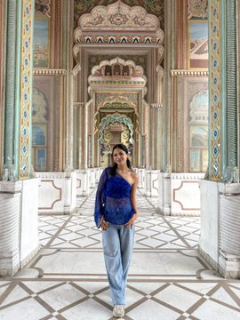 A woman in a blue off-the-shoulder top and jeans poses in the center of an ornate, palatial hallway with decorative columns and patterned floors.
