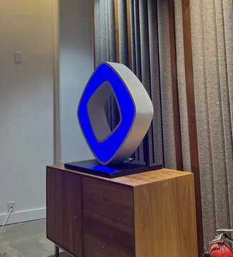 A modern square-shaped blue illuminated lamp sitting on a wooden cabinet next to a window with curtains.