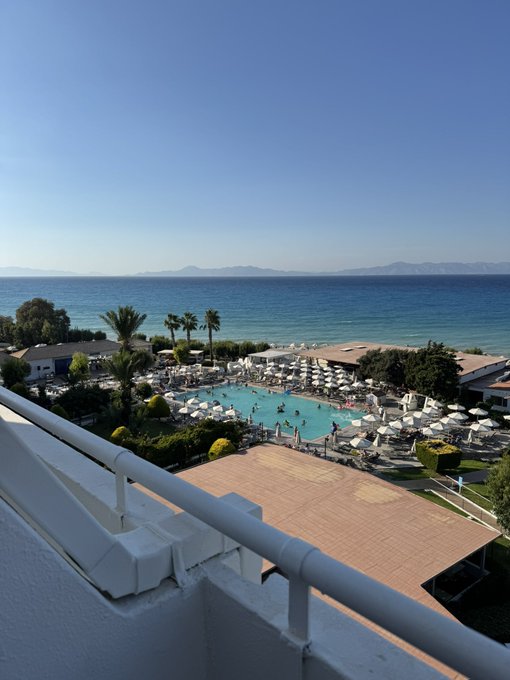A scenic view from a hotel balcony overlooking a beach resort with a large swimming pool, white umbrellas, palm trees, and the calm sea stretching toward distant mountains under a clear blue sky.