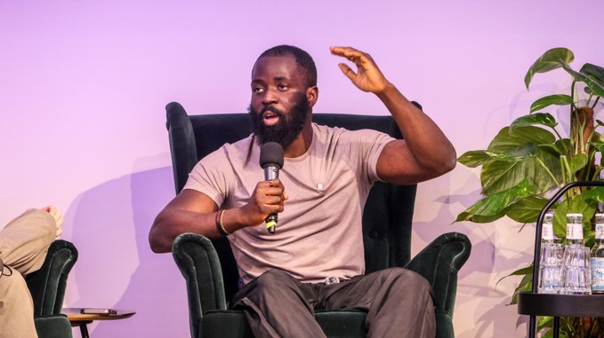 A bearded man in a tan t-shirt speaks into a microphone while seated in a green velvet armchair, gesturing with his left hand at what appears to be a panel discussion.
