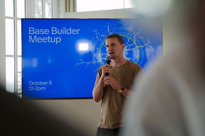 A man speaks into a microphone at a 'Base Builder Meetup' event on October 8, 12-2pm, standing in front of a blue screen.