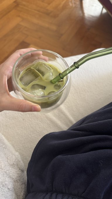 A first-person view of a hand holding a clear glass cup of iced matcha with a green glass straw, while the person sits comfortably under a white blanket.