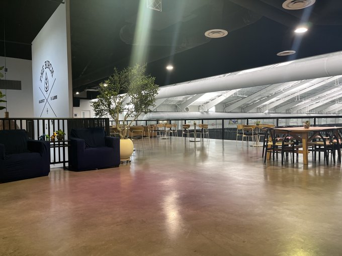 An interior view of a modern lounge or cafe with polished concrete floors, armchairs, potted plants, and dining tables under industrial ceilings with exposed pipes.