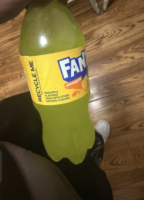 A hand holds a bottle of Pineapple Fanta (FANta) saying "Pineapple Flavored Soda with Other Natural Flavors" and "Recycle Me - PEEL PUSH ENJOIN ENJOY" above a wooden floor with white sneakers visible in the background.