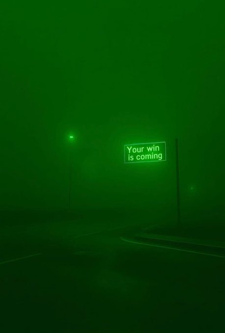 A foggy nighttime street scene illuminated in green, featuring a glowing sign that reads 'Your win is coming' next to a streetlight.