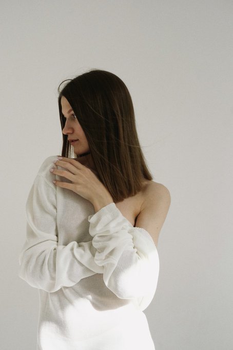 A side profile of a young woman with shoulder-length brown hair wearing a white off-the-shoulder top, posing against a plain light background.