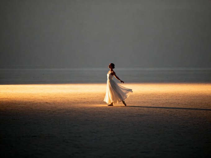 A woman in a flowing white dress stands alone in a vast, empty landscape with a strip of golden light illuminating the ground around her.