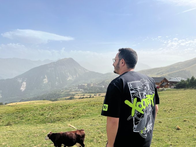 A man wearing sunglasses and a black t-shirt featuring 'XCOPY' artwork stands in a grassy alpine meadow with mountains in the background while a brown and white dog runs by.