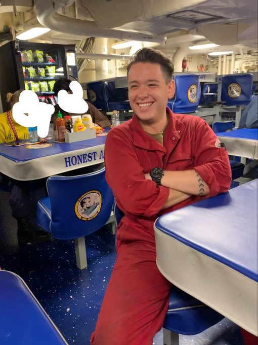 A smiling young man in a red coverall with arms crossed sits at a table inside what appears to be a naval vessel's mess hall; a table labeled 'HONEST A...' is visible behind him.