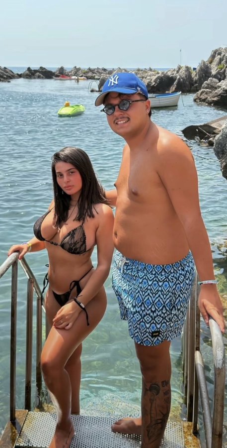 A couple stands on dock stairs by clear turquoise water, with the man wearing a blue NY Yankees cap and sunglasses, while the post "Remember you're not ugly, you just don't hold 1% TBB" humorously implies confidence and attractiveness, possibly playing on the man's self-assured pose.