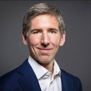 A headshot portrait of Bitwise CIO Matt Hougan, a man with short graying hair wearing a dark blue blazer over a light blue shirt, smiling at the camera against a gray background. The image accompanies coverage of his bullish outlook on blockchain adoption, where he predicts massive growth in on-chain transaction volume driven by tokenization and AI-powered trading.