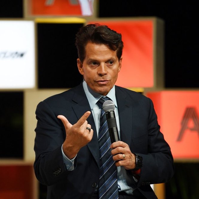 A man in a dark suit and striped tie speaking into a microphone and gesturing with his hand, standing in front of a backdrop featuring orange and yellow squares.