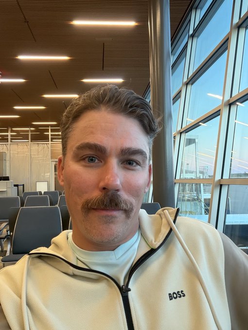 A selfie of a young man with a mustache wearing a cream-colored BOSS hoodie, taken in what appears to be an airport waiting area with seating and large windows.
