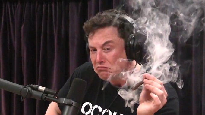 A man wearing headphones and a black t-shirt sits in front of a microphone, smoking a cigar with visible smoke swirling around his face.