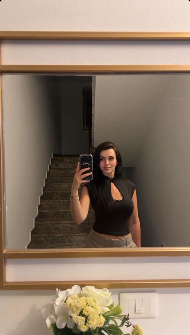 A mirror selfie shows a woman in a black halter top and beige pants standing on a staircase, with a vase of white flowers visible below the mirror.