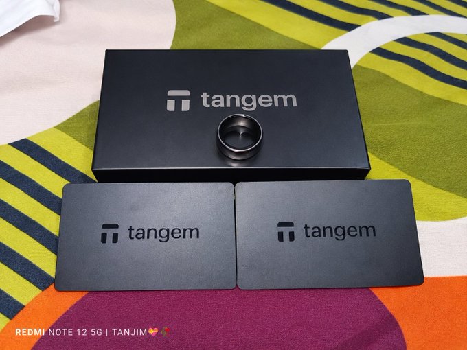The image shows a Tangem hardware wallet set, featuring a black box labeled "tangem" with a ring-shaped device on top and two "tangem" card-style wallets displayed in front, all arranged on a colorful patterned cloth. The photo illustrates the post's message about receiving a secure, user-friendly self-custody solution for crypto storage.