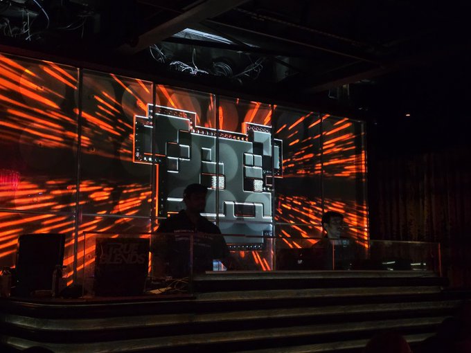 A DJ performs on stage at 'True Blends,' with a large pixel-art graphic projected on screens behind an orange, light-streaked backdrop.