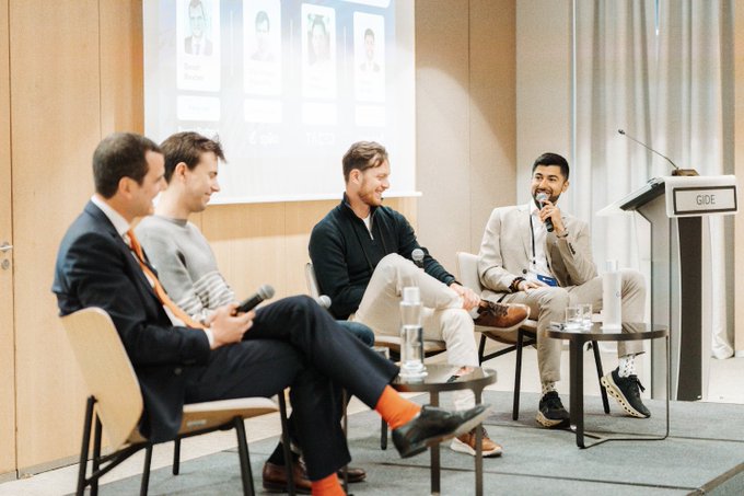 Four men in business attire participate in a panel discussion at a CIBE event, with one man speaking into a microphone while others listen and hold microphones, with a projected slide showing speaker profiles behind them.