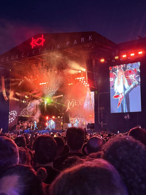 The image shows a large crowd of people gathered at an outdoor concert, with many of them standing close together. The stage features a large screen displaying a video or photo of a woman's face, possibly a performer or a promotional image related to the event. The audience appears engaged and excited about the performance, creating a lively atmosphere.