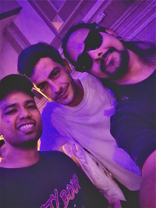 Three friends pose for a selfie under purple-lit lighting at what appears to be a lively event, with the caption suggesting they're in Singapore for Token2049. The person on the right wears sunglasses and a slight smirk, while the man in the middle sports a white shirt with visible stains, and the smiling man on the left wears a cap and a shirt with "NY BOY" partially visible.