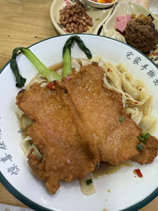 A close-up photo of a Chinese dish featuring a large fried meat cutlet served over wide noodles in broth, garnished with greens, alongside small side dishes including peanuts and meatballs.