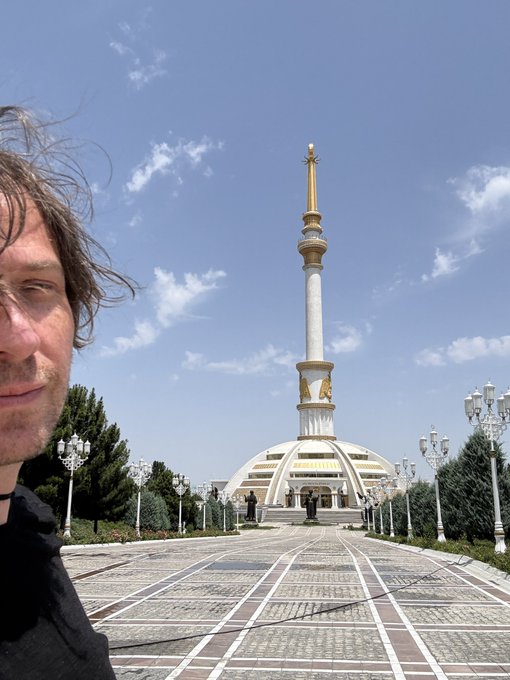 The image features a man standing next to a tall white monument with gold accents, possibly a temple or a significant building. The man appears to be posing for a photo while looking at the camera. The background of the image shows trees and other buildings, indicating that this scene takes place in an urban setting. The caption provided seems unrelated to the main subject matter but is likely meant to provide context or additional information about the monument or the man's location.