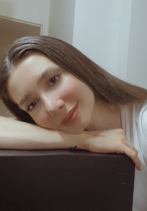 The image features a woman with long brown hair leaning against a black couch, smiling at the camera. The background of the photo appears to be white, which contrasts nicely with her dark hair. There are no texts or other objects present in the image, making it a simple and uncluttered composition that highlights the woman's face and expression.