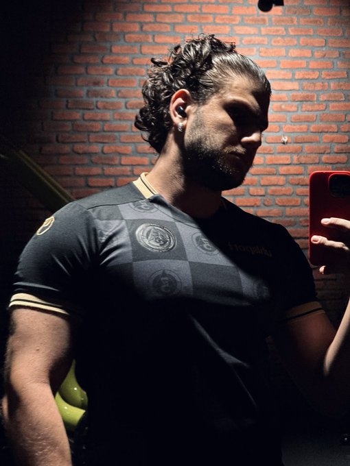 A muscular man with curly hair in a black patterned t-shirt takes a mirror selfie with a red phone against a brick wall.