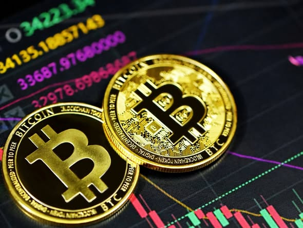 Two physical Bitcoin tokens rest on a financial chart with candlestick patterns and visible numbers including '3A221', '54135.18057143', '33687.97600000', and '22978.69868868'.