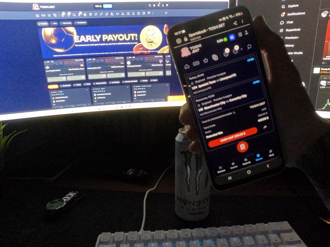 A hand holds a smartphone displaying the "Toshibet - TOOSE" app with an "EARLY PAYOUT" promotion and a red "Claim KSH 2,863" button, alongside a Monster Energy drink can in front of a laptop showing the same betting interface. The scene directly illustrates the post's excitement about gambling on Toshibet while enjoying the Monster energy drink.