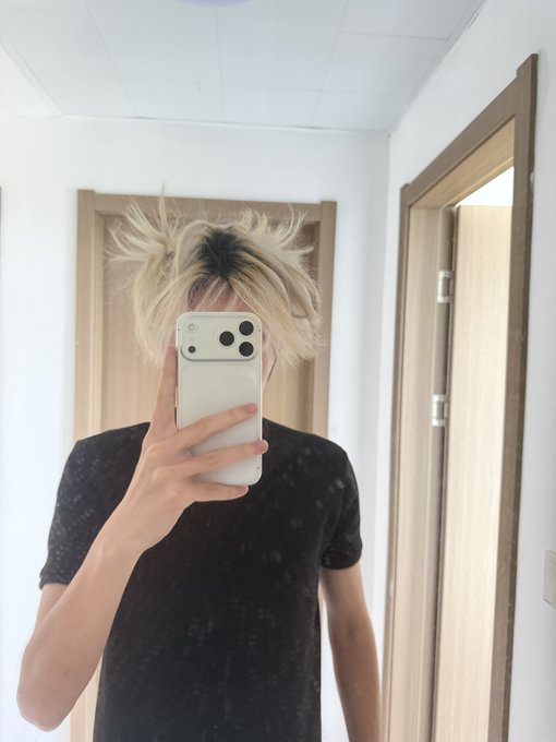 Mirror selfie of a person with messy blonde hair taking a photo with an iPhone, wearing a black t-shirt in a white hallway.
