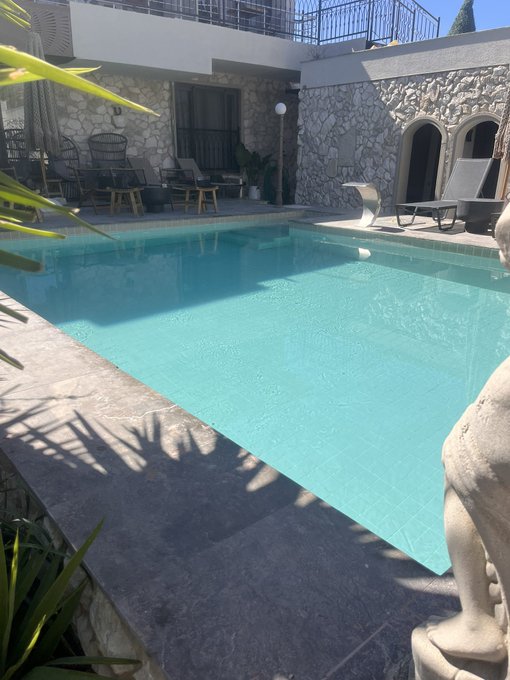 Outdoor courtyard scene featuring a rectangular swimming pool with clear turquoise water, surrounded by a stone-walled building with arched doorways, patio furniture, and lounge chairs under bright sunlight.