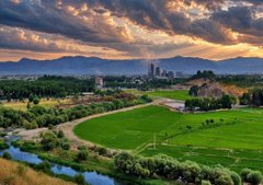 A blurred landscape photograph showing a winding river, green fields, and trees beneath a dramatic sunset sky with orange and grey clouds.