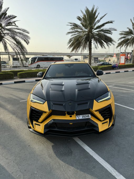A striking yellow and black Lamborghini Urus with a Dubai license plate reading "U 39340" sits parked in a lot lined with palm trees, ready to greet followers with a luxurious "Good Morning GOATS."